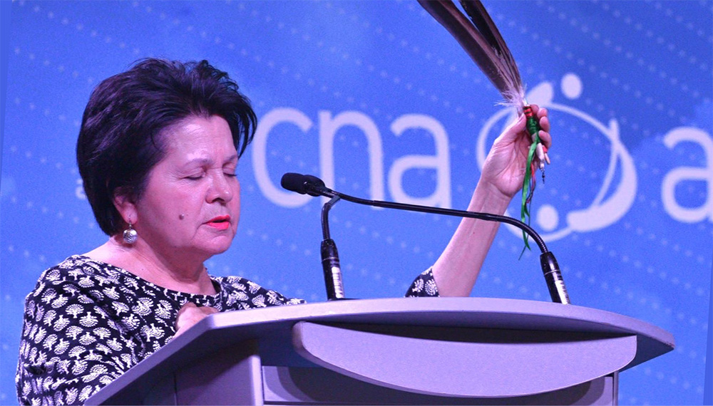 A woman standing at a podium, speaking into microphones with her eyes closed, as if in a moment of reflection or dedication. She holds a feather aloft with her right hand, symbolizing respect, honor, or a cultural or spiritual gesture